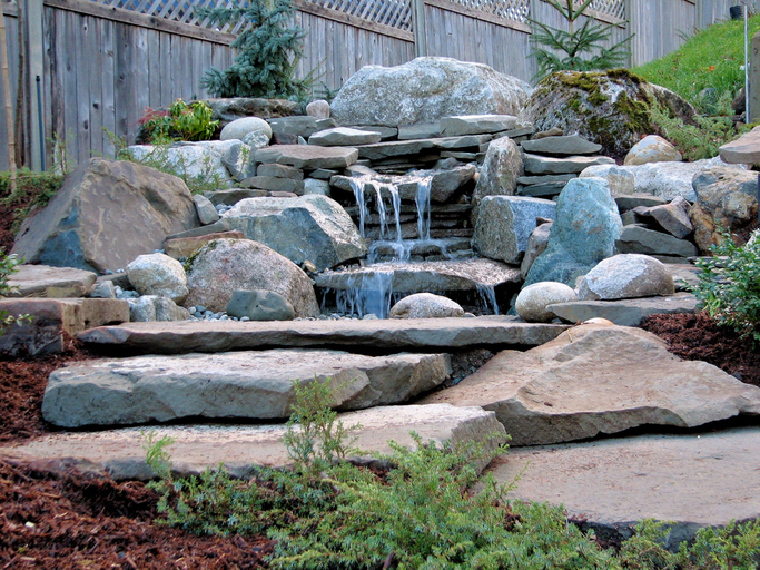 A garden waterfall built with mostly slabs of sandstone and surrounded by some granite boulder - rocks carefully placed into the existing hillside with flowing water.